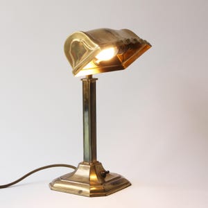 Op de afbeelding: Een vintage messing bureaulamp met een gebogen kap en een rechthoekige voet. De lamp heeft een warme, gouden tint en een klassiek ontwerp. De gloeilamp is zichtbaar en verlicht de omgeving. De lamp is aangesloten op een bruin elektrisch snoer.