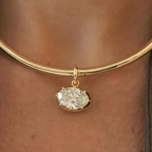 May include: A gold necklace featuring an oval diamond pendant. The necklace has a smooth, rounded band. The diamond pendant is set in a gold bezel and hangs from a small loop. The necklace is a simple, elegant design.