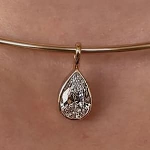 Puede incluir: Un collar dorado con un colgante en forma de lágrima con una piedra preciosa clara y brillante. El colgante cuelga de una delicada cadena circular dorada. La joya se muestra sobre un fondo color piel.