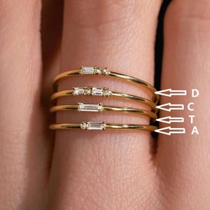 Puede incluir: Cuatro anillos de oro, cada uno con un solo diamante baguette. Los anillos están apilados, con el más grande en la parte inferior y el más pequeño en la parte superior. Los anillos están etiquetados como A, T, C y D de abajo hacia arriba.