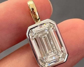 May include: A rectangular emerald-cut diamond pendant with a gold bail. The diamond is set in a silver bezel. The pendant is held in a person's hand against a gray background.