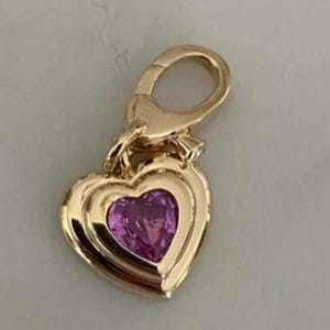 Può includere: Un ciondolo a forma di cuore color oro con una pietra preziosa rosa sfaccettata al centro. Il ciondolo include una chiusura a moschettone dorata sulla parte superiore, che consente di agganciarlo a una catenina o a un braccialetto.