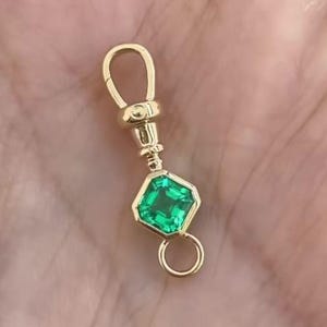 Può includere: Una chiusura girevole in oro con una pietra preziosa verde a taglio quadrato incastonata in una montatura d'oro. La chiusura presenta un occhiello arrotondato sulla parte superiore e un anello circolare più piccolo per fissare gioielli.