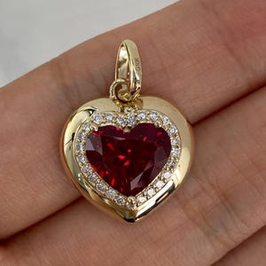 Puede incluir: Un colgante en forma de corazón de oro de 10K con una gran piedra roja facetada en el centro, rodeada por un halo de pequeñas piedras claras. El asa de metal en la parte superior tiene grabado el texto 10K, indicando la pureza del oro.