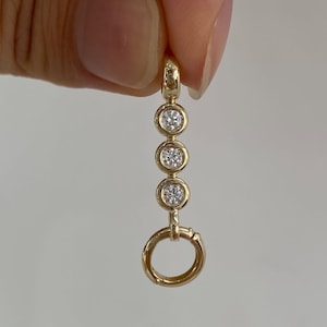 Puede incluir: Un colgante de oro con una fila vertical de tres piedras redondas, claras y facetadas, engastadas en biseles de oro pulido, rematado con una pequeña anilla de oro y terminado con un aro de oro en la parte inferior, de diseño lineal.