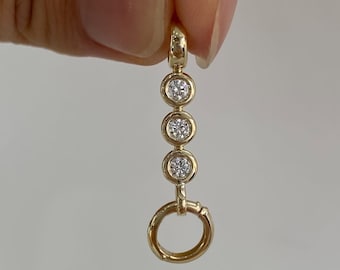 May include: A gold pendant featuring a vertical row of three round, bezel-set clear stones, topped with a small circular bail and finished with a larger, plain gold circular ring at the bottom, creating a linear, multi-component jewelry piece.