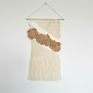 May include: A beige and white woven wall hanging with a fringe. The hanging features a geometric pattern with brown flower accents.