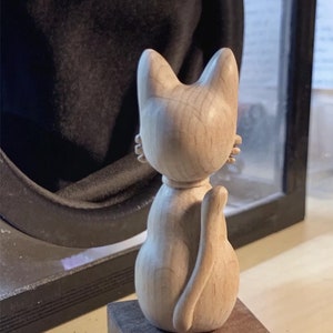 Puede incluir: Una figurita de gato de madera de color marrón claro. El gato está sentado en un pequeño bloque de madera. El gato está de espaldas a la cámara, con la cola enroscada alrededor de su cuerpo.
