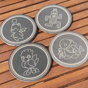 Puede incluir: Cuatro posavasos redondos de piedra gris oscuro con diseños grabados en blanco. Cada posavasos presenta un personaje diferente de un videojuego. Los posavasos están dispuestos sobre una superficie de madera.