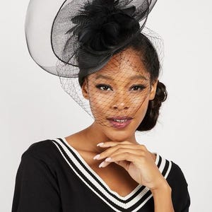 May include: A black hat with a veil, adorned with a cluster of black feathers. The hat features a wide brim and a net veil that covers the face. The woman is wearing a black sweater with white stripes.