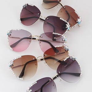 May include: An assortment of rimless sunglasses with gradient lenses in various shades of brown, pink, and purple. The frames are gold-toned, and the edges are embellished with rhinestones and small floral accents.