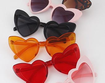 May include: A collection of heart-shaped sunglasses in various colors, including pink, black, red, and orange. Some frames have a solid color, while others feature a tortoiseshell pattern. The sunglasses are arranged on a white surface.