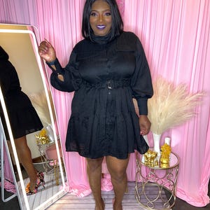 May include: A black dress with long sleeves, a button-down front, and a tiered skirt. The dress has a belt at the waist and is paired with colorful patterned heels. The background features a pink curtain and a gold side table.