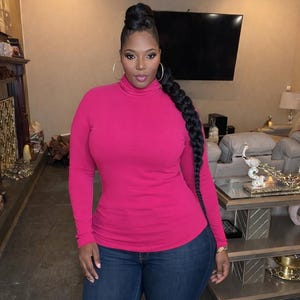 May include: A woman wearing a long-sleeved, fuchsia turtleneck top and blue jeans. She has large hoop earrings and a long, braided hairstyle. The background shows a living room with a fireplace, a large TV, and a sofa.