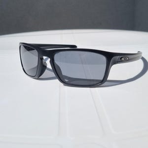 Puede incluir: Gafas de sol negras con lentes grises. Las gafas tienen una montura rectangular y una patilla curva. El logotipo de Oakley es visible en la patilla. Las gafas est&aacute;n sobre una superficie blanca.