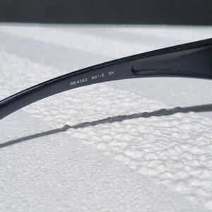 May include: A black plastic temple arm of a pair of sunglasses. The interior surface of the arm features white printed text that reads RB 4093 601-S 3N. The arm has a smooth, curved design and is positioned against a textured white surface.