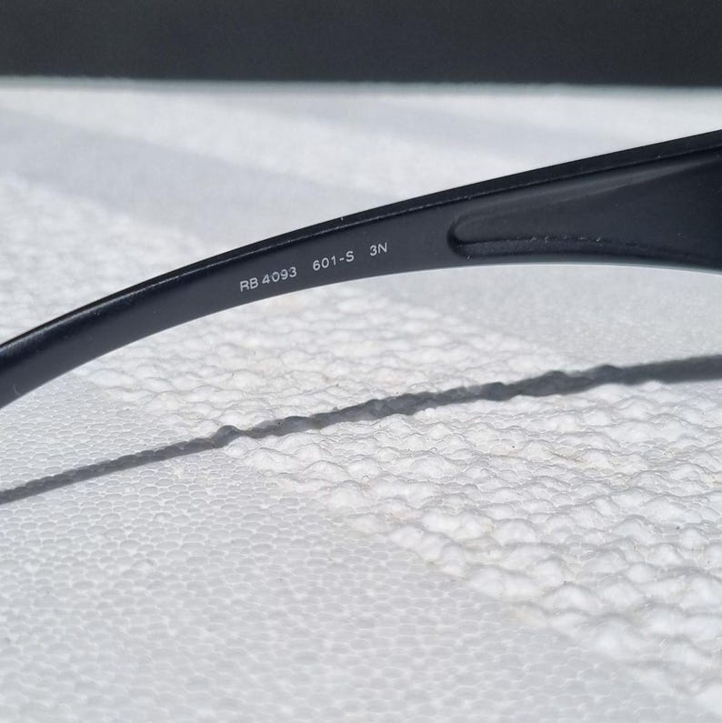 May include: A black plastic temple arm of a pair of sunglasses. The interior surface of the arm features white printed text that reads RB 4093 601-S 3N. The arm has a smooth, curved design and is positioned against a textured white surface.