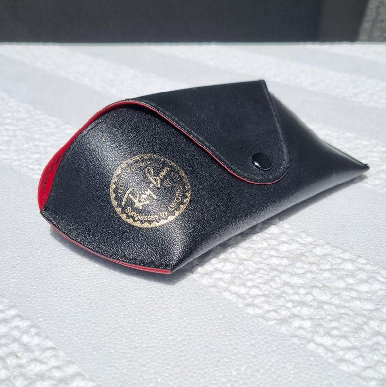 May include: A black, envelope-style sunglasses case with a red interior lining and a snap button closure. The front features a circular gold-toned emblem with the text Ray-Ban Sunglasses by Luxottica printed in black, centered on the flap.