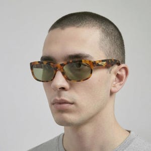 May include: Sunglasses with a tortoiseshell pattern and green lenses. The frames are rectangular, with a small gold emblem on the temples. The sunglasses are worn by a person.