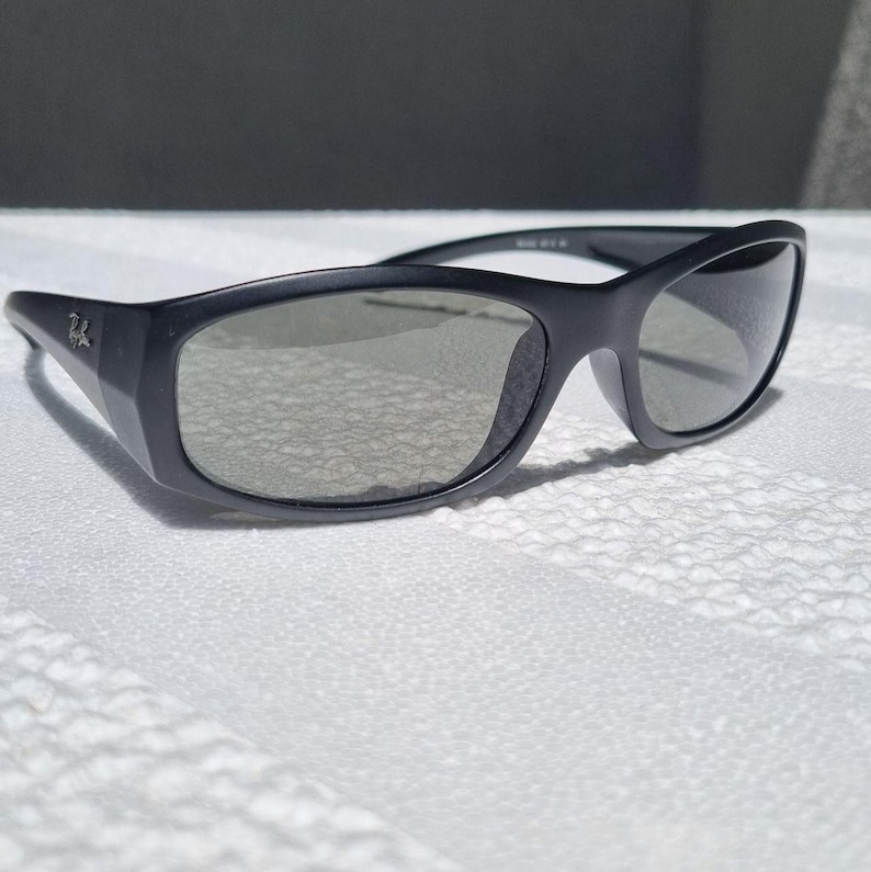 May include: Black sunglasses with a matte finish and dark grey lenses. The frames feature a wrap-around design with a small silver logo on the temple. The lenses are semi-transparent, showing a subtle reflection of the surrounding environment.