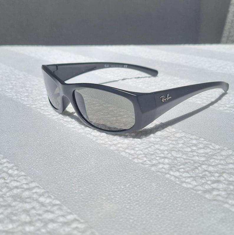 May include: Dark grey sunglasses with a wraparound frame design and tinted lenses. The Ray-Ban logo is printed in white on the outer temple of the frame. These sunglasses feature a smooth, solid-colored finish and a curved shape for coverage.
