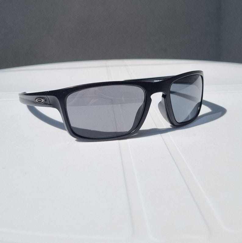 Puede incluir: Gafas de sol negras con montura rectangular y lentes grises. Las gafas de sol est&aacute;n sobre una superficie blanca. El logotipo de Oakley es visible en la patilla.
