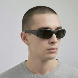 May include: Black wrap-around sunglasses with dark tinted lenses and a matte finish frame. The temples feature the Ray-Ban logo in small white lettering. The design provides full coverage around the eyes with a curved, aerodynamic shape.