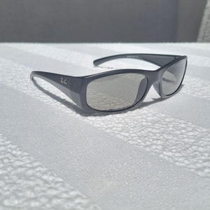 May include: Dark gray sunglasses with a wrap-around frame design and light gray tinted lenses. The temples feature the Ray-Ban logo in silver lettering. These sunglasses have a smooth, matte finish and a curved shape that contours to the face.