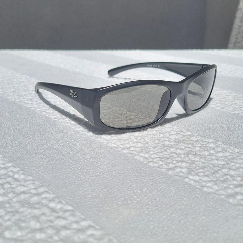 May include: Dark gray sunglasses with a wrap-around frame design and light gray tinted lenses. The temples feature the Ray-Ban logo in silver lettering. These sunglasses have a smooth, matte finish and a curved shape that contours to the face.