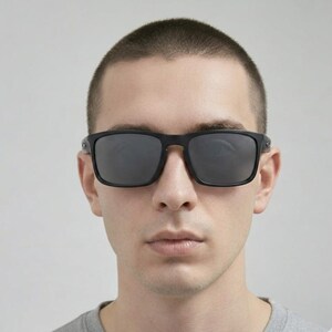 Puede incluir: Gafas de sol negras con montura cuadrada y lentes oscuras. Las gafas de sol tienen un acabado mate y las lleva una persona. La imagen es un primer plano de la cara, centr&aacute;ndose en las gafas de sol.