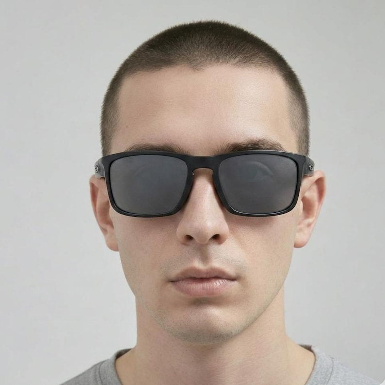 Puede incluir: Gafas de sol negras con montura cuadrada y lentes oscuras. Las gafas de sol tienen un acabado mate y las lleva una persona. La imagen es un primer plano de la cara, centr&aacute;ndose en las gafas de sol.