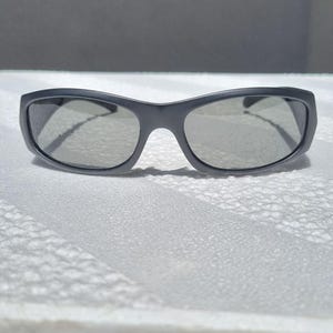 May include: A pair of matte charcoal gray sunglasses featuring a wraparound frame design with dark tinted lenses. The eyewear has a smooth, solid-colored plastic construction with a curved bridge and wide temples that extend from the outer corners.