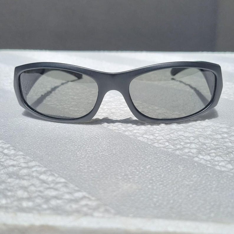 May include: A pair of matte charcoal gray sunglasses featuring a wraparound frame design with dark tinted lenses. The eyewear has a smooth, solid-colored plastic construction with a curved bridge and wide temples that extend from the outer corners.