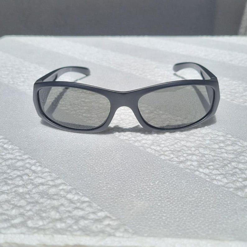 May include: A pair of matte charcoal gray sunglasses featuring a wraparound frame design with dark tinted lenses. The eyewear has a curved bridge and thick temples that extend from the outer corners of the frame, providing a sleek look.