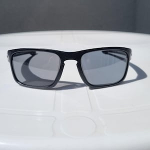 Puede incluir: Gafas de sol negras con lentes grises. Las gafas tienen una montura rectangular y est&aacute;n sobre una superficie blanca. Las lentes son reflectantes y proyectan una sombra.