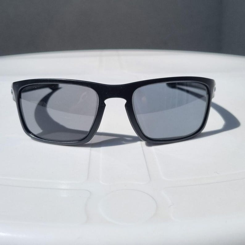 Puede incluir: Gafas de sol negras con lentes grises. Las gafas tienen una montura rectangular y est&aacute;n sobre una superficie blanca. Las lentes son reflectantes y proyectan una sombra.