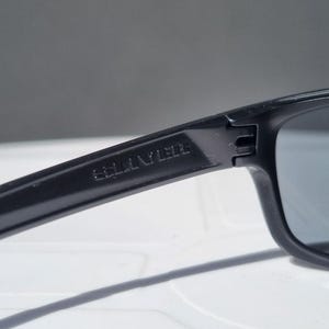 Puede incluir: Primer plano de gafas de sol negras con el nombre de la marca "SELLINYLEDE" grabado en la patilla. Las gafas tienen una montura rectangular y lentes oscuras, adecuadas para la protecci&oacute;n solar.