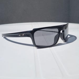 Puede incluir: Gafas de sol negras con montura rectangular y lentes grises. Las gafas de sol tienen un dise&ntilde;o elegante y moderno con un logotipo sutil en la patilla. Las gafas de sol est&aacute;n sobre una superficie blanca.