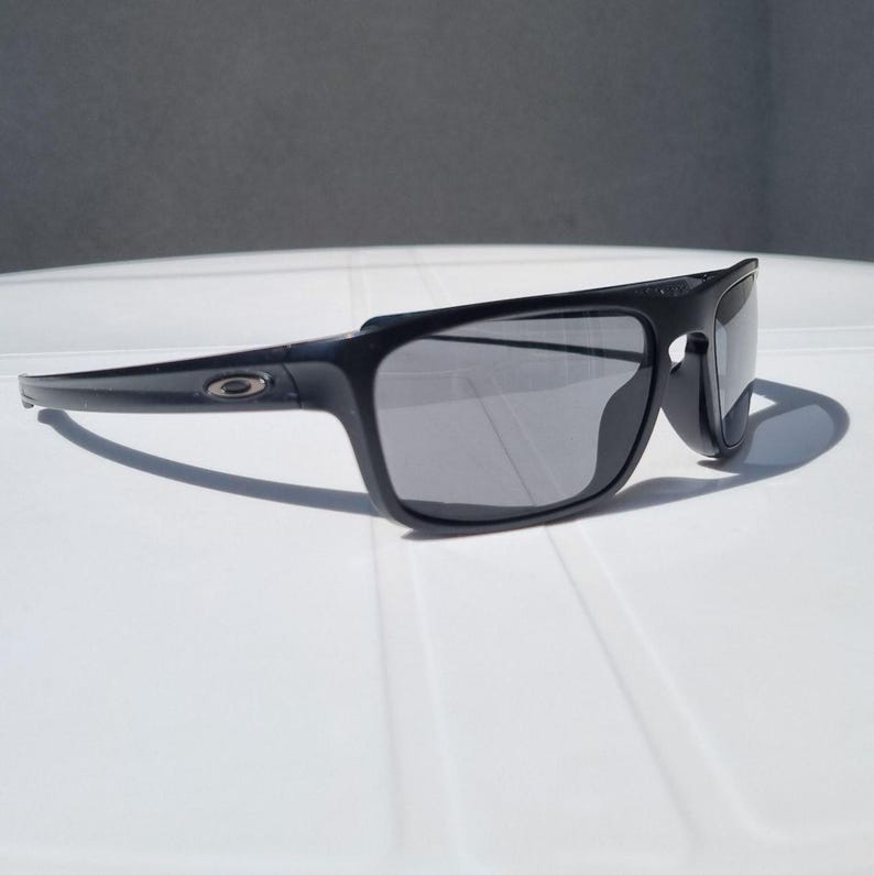 Puede incluir: Gafas de sol negras con montura rectangular y lentes grises. Las gafas de sol tienen un dise&ntilde;o elegante y moderno con un logotipo sutil en la patilla. Las gafas de sol est&aacute;n sobre una superficie blanca.