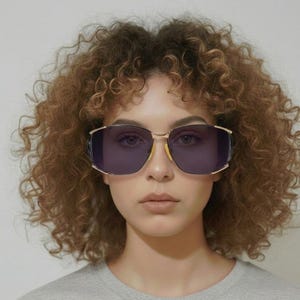 May include: Gold-framed sunglasses with dark purple lenses. The sunglasses have a rectangular shape with a slight cat-eye design. The frame is gold-colored, and the lenses are a deep purple. The sunglasses are worn by a person with curly brown hair.