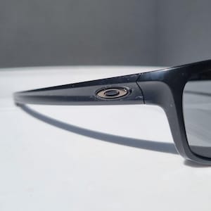 Puede incluir: Primer plano de gafas de sol negras con lentes gris oscuro. Las gafas tienen una montura rectangular y un peque&ntilde;o logotipo circular en la patilla. La montura es de material negro brillante.
