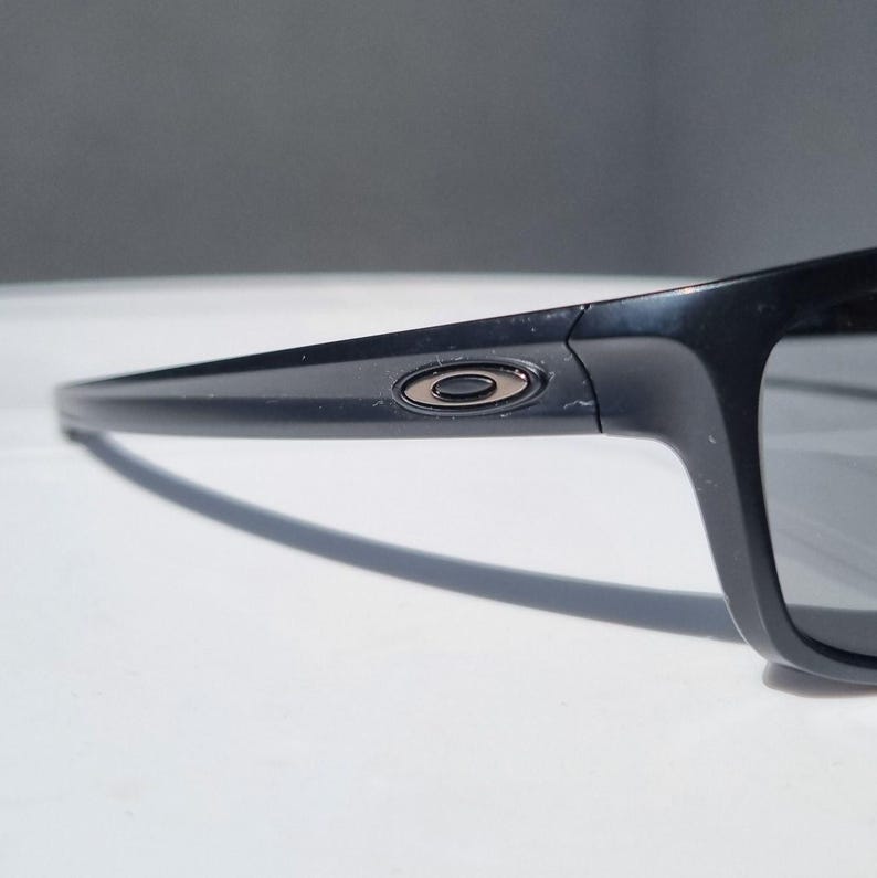 Puede incluir: Primer plano de gafas de sol negras con lentes gris oscuro. Las gafas tienen una montura rectangular y un peque&ntilde;o logotipo circular en la patilla. La montura es de material negro brillante.