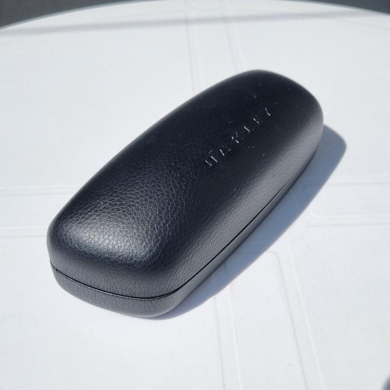 Puede incluir: Un estuche negro y rectangular para gafas con el nombre de la marca "OAKLEY" grabado en la parte superior. El estuche tiene una superficie texturizada y una forma redondeada. El estuche est&aacute; cerrado y parece estar hecho de un material duradero.
