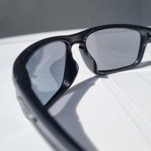 Puede incluir: Gafas de sol negras con lentes grises. Las gafas tienen una montura gruesa y rectangular y un dise&ntilde;o curvo. Las lentes son reflectantes y las gafas descansan sobre una superficie blanca.