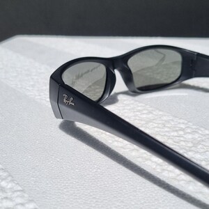 May include: Black sunglasses with a matte finish and dark tinted lenses. The frame features a wrap-around design with thick temples. The Ray-Ban logo is printed in white on the outer side of the left temple near the hinge, clearly visible.