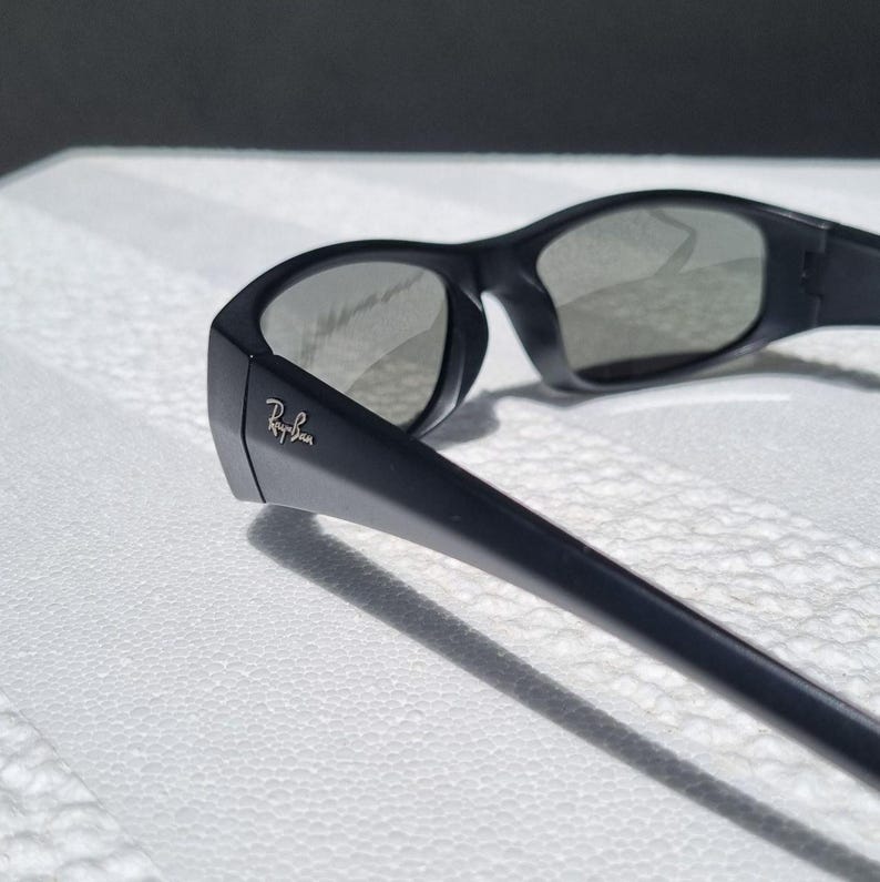 May include: Black sunglasses with a matte finish and dark tinted lenses. The frame features a wrap-around design with thick temples. The Ray-Ban logo is printed in white on the outer side of the left temple near the hinge, clearly visible.