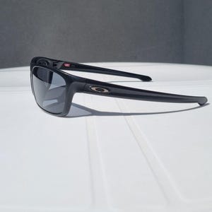 Puede incluir: Gafas de sol negras con montura rectangular y lentes oscuras. Las gafas de sol tienen un dise&ntilde;o elegante con el logotipo de Oakley en las patillas. La montura est&aacute; hecha de un material duradero y las lentes protegen del sol.