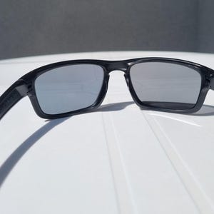 Puede incluir: Gafas de sol negras con montura rectangular y lentes grises. Dise&ntilde;o elegante y moderno, con acabado mate en la montura. Las lentes son reflectantes y las patillas ligeramente curvadas. Adecuadas para actividades al aire libre.