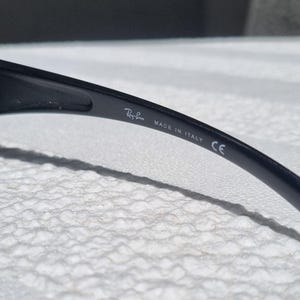 May include: A black temple arm of a pair of sunglasses featuring the white printed text Ray-Ban, MADE IN ITALY, and the CE mark. The smooth, curved plastic surface shows the brand branding clearly on the inner side of the frame component.