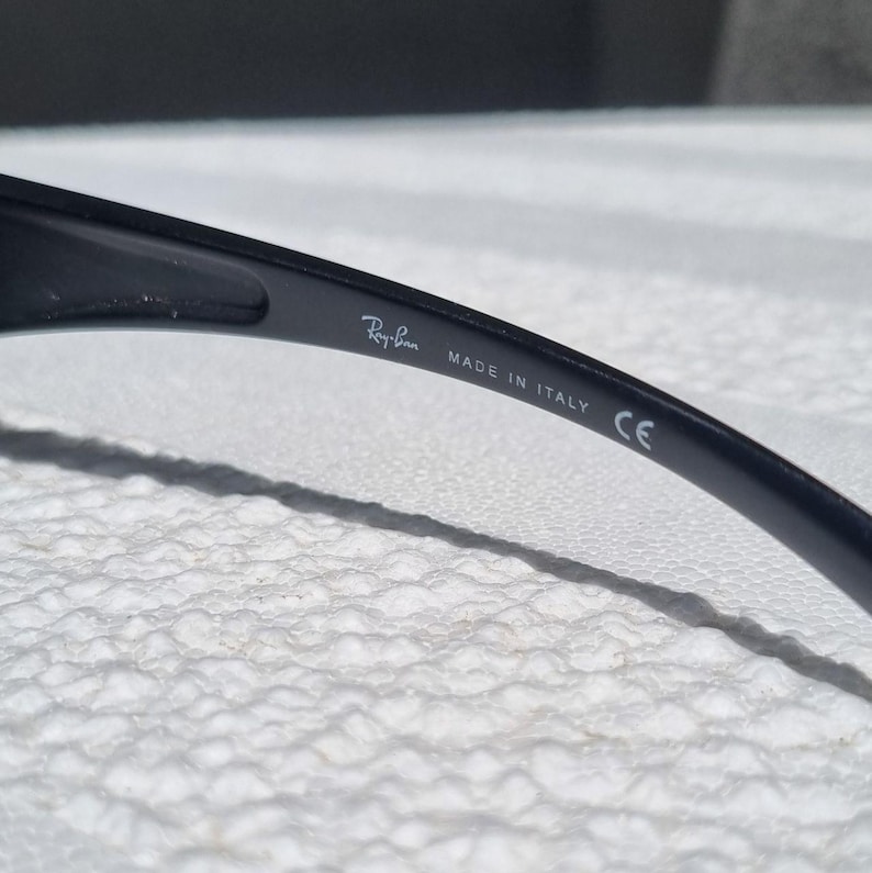 May include: A black temple arm of a pair of sunglasses featuring the white printed text Ray-Ban, MADE IN ITALY, and the CE mark. The smooth, curved plastic surface shows the brand branding clearly on the inner side of the frame component.